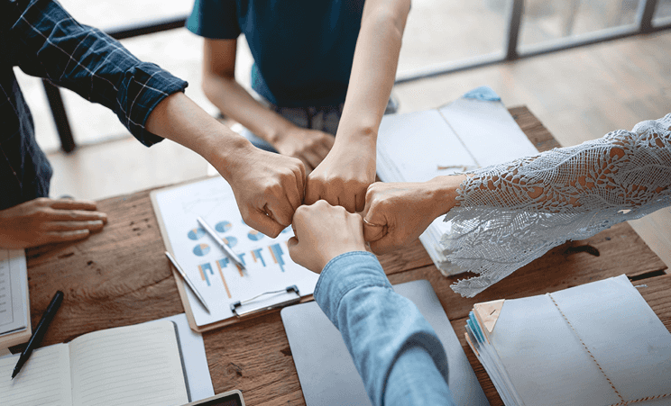 4 people bumping fists together over documents with charts and graphs on them